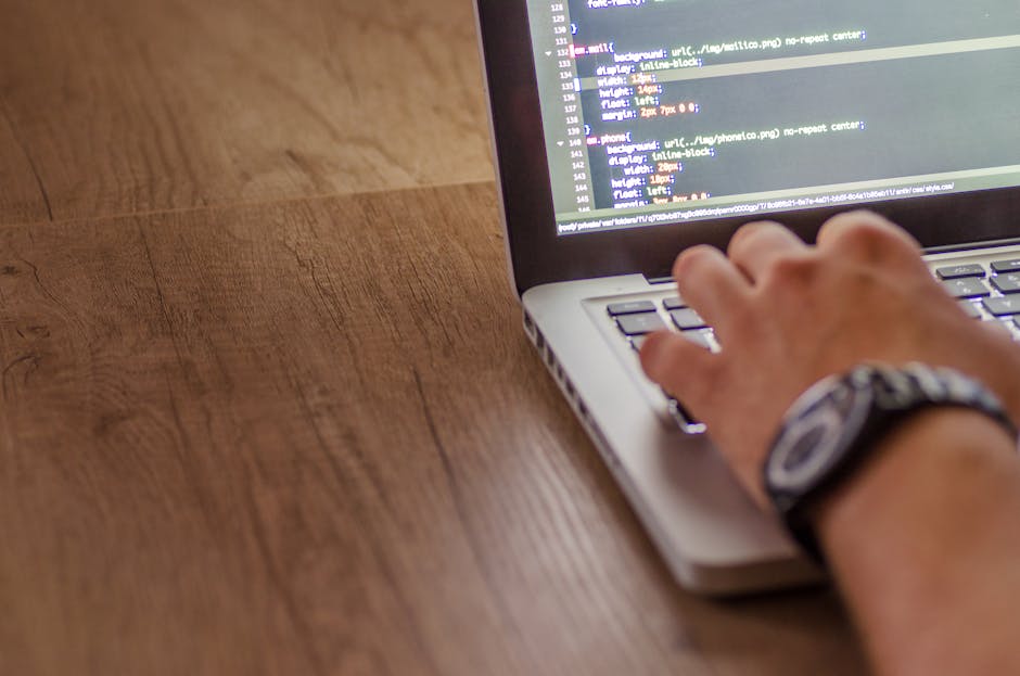 A person's hand using a laptop computer on a wooden surface, with the screen displaying lines of software code. The individual is wearing a wristwatch and appears to be engaged in tasks related to dig